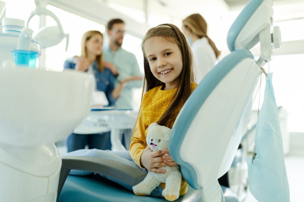child sitting in dental chair smiling and holding plush bear